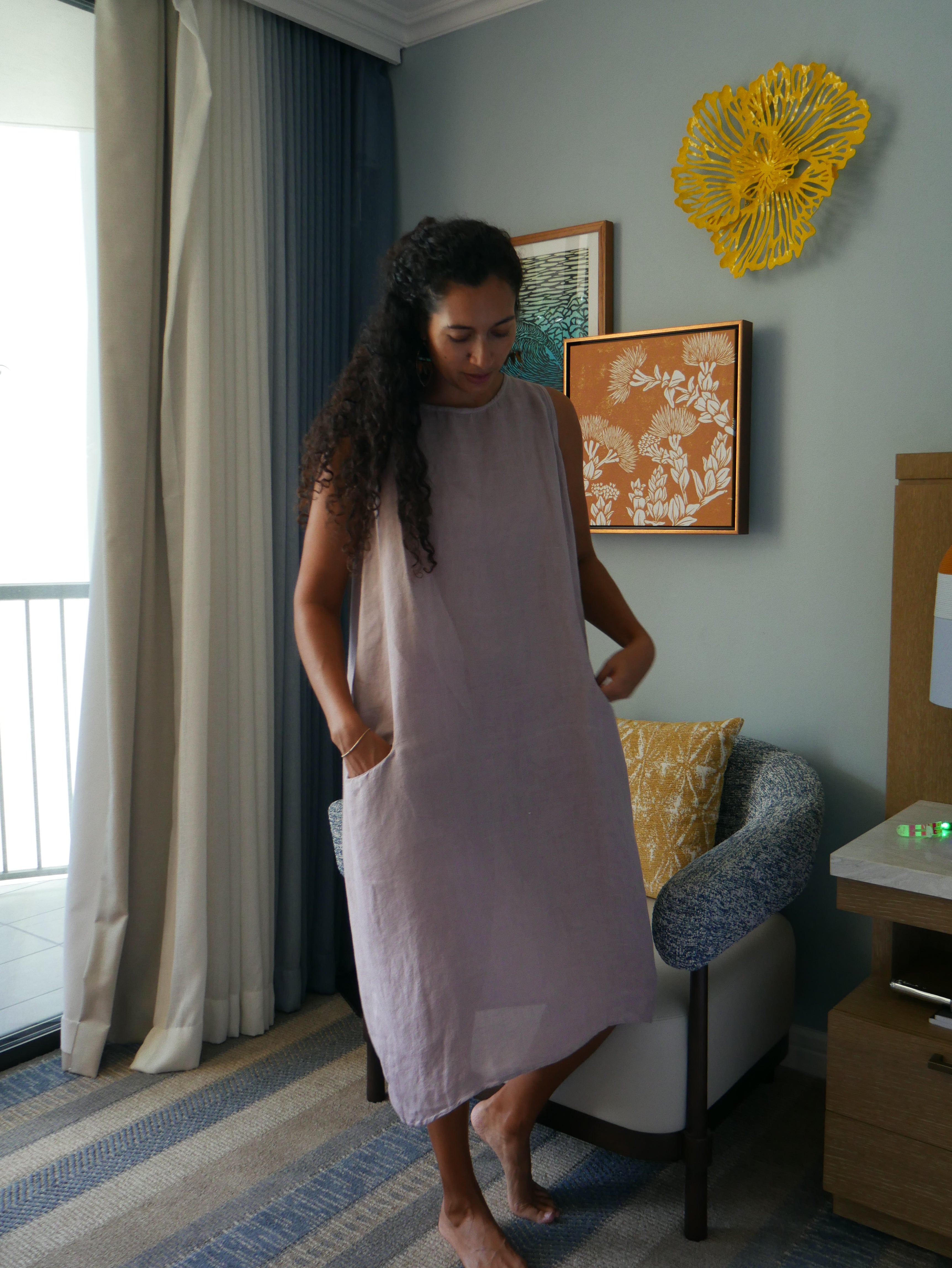 Model in a soft Kahoʻolawe-toned sleeveless linen midi dress with round neckline and pockets, wearing lei, photographed on a Hilo farm.