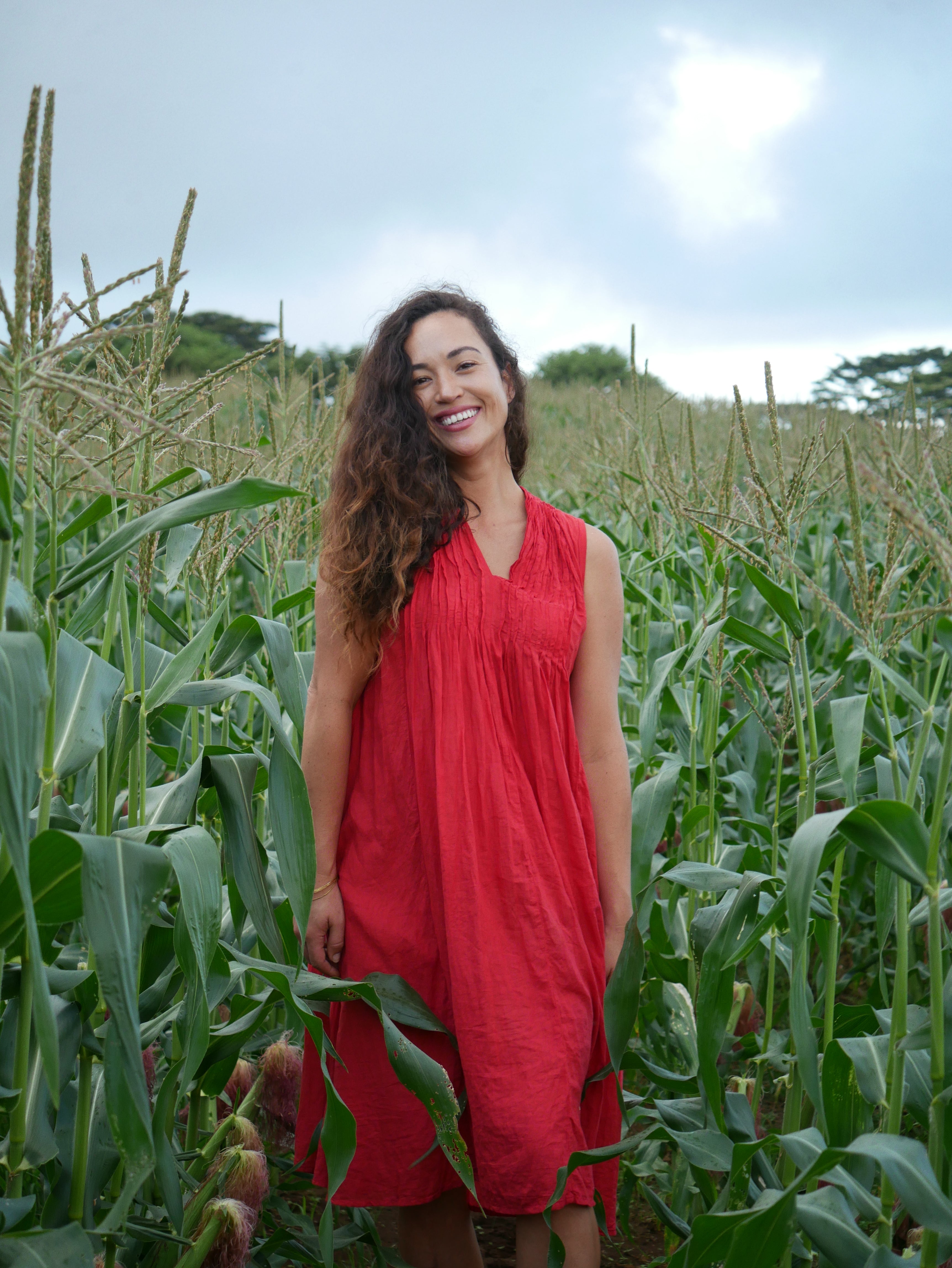 Model in a deep lychee-red cotton A-line dress with V-neckline and pockets, photographed on a Hilo farm.