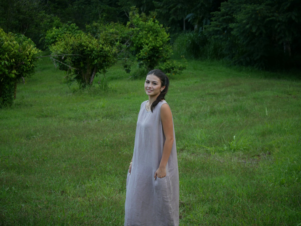 Model in a soft Kahoʻolawe-toned sleeveless linen midi dress with round neckline and pockets, wearing lei, photographed on a Hilo farm.
