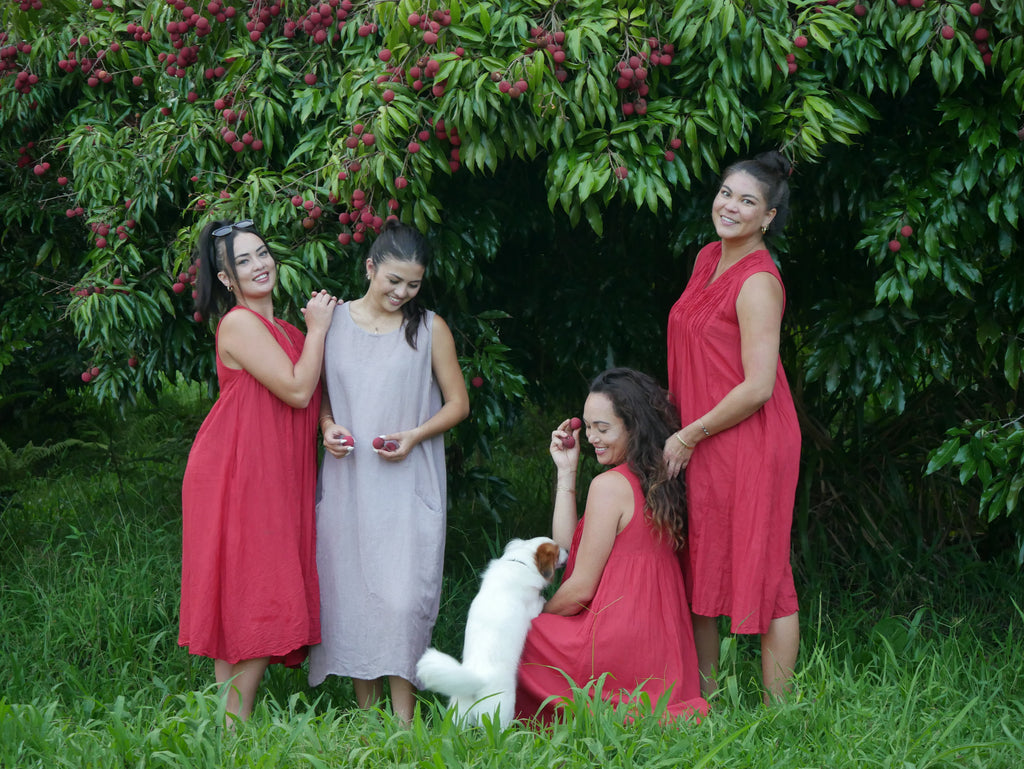 Model in a deep lychee-red cotton A-line dress with V-neckline and pockets, photographed on a Hilo farm.