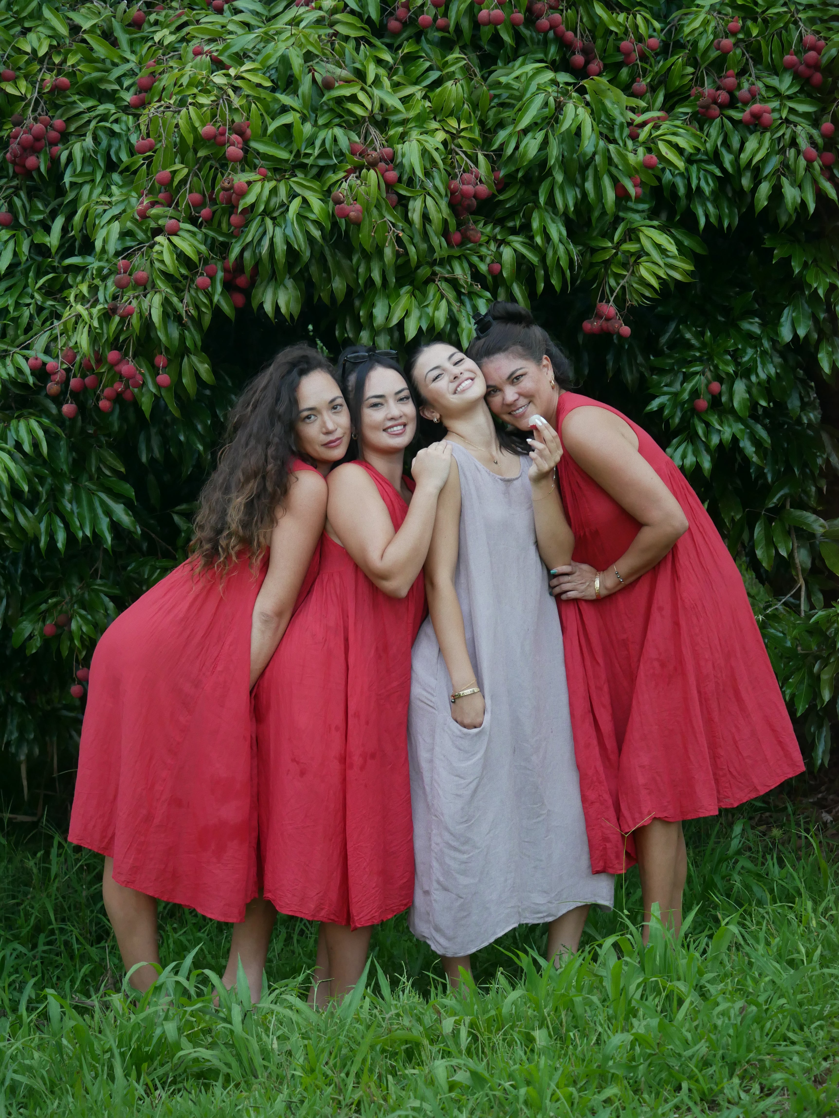 Model in a deep lychee-red cotton A-line dress with V-neckline and pockets, photographed on a Hilo farm.