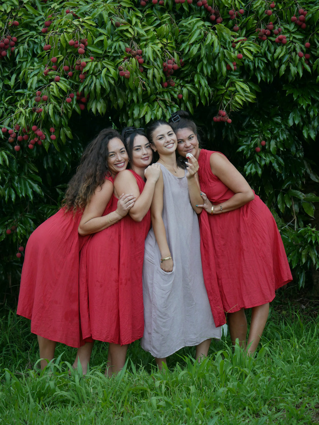 Model in a deep lychee-red cotton A-line dress with V-neckline and pockets, photographed on a Hilo farm.
