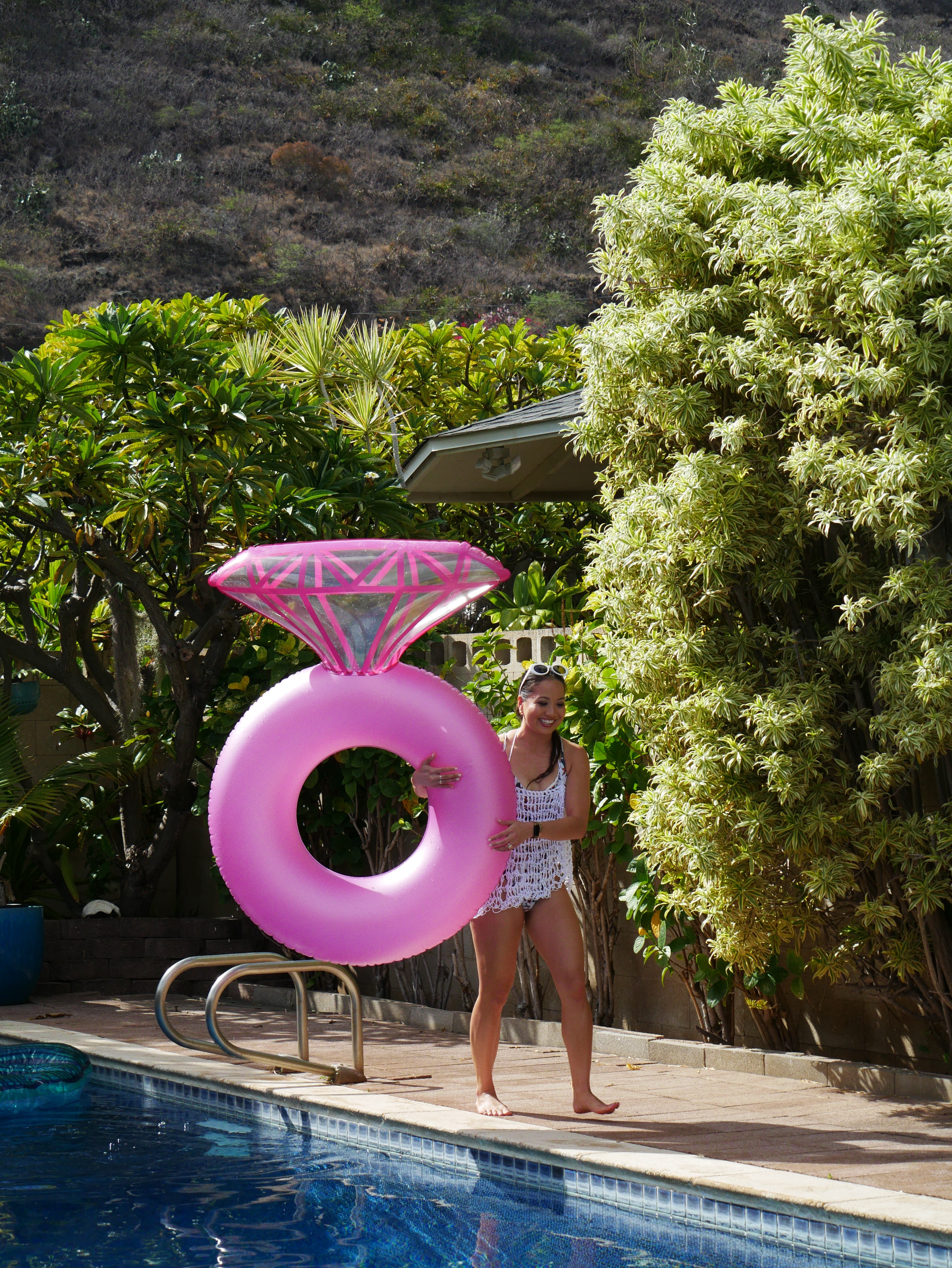 Person holding a pink inflatable ring by a pool with trees in the background